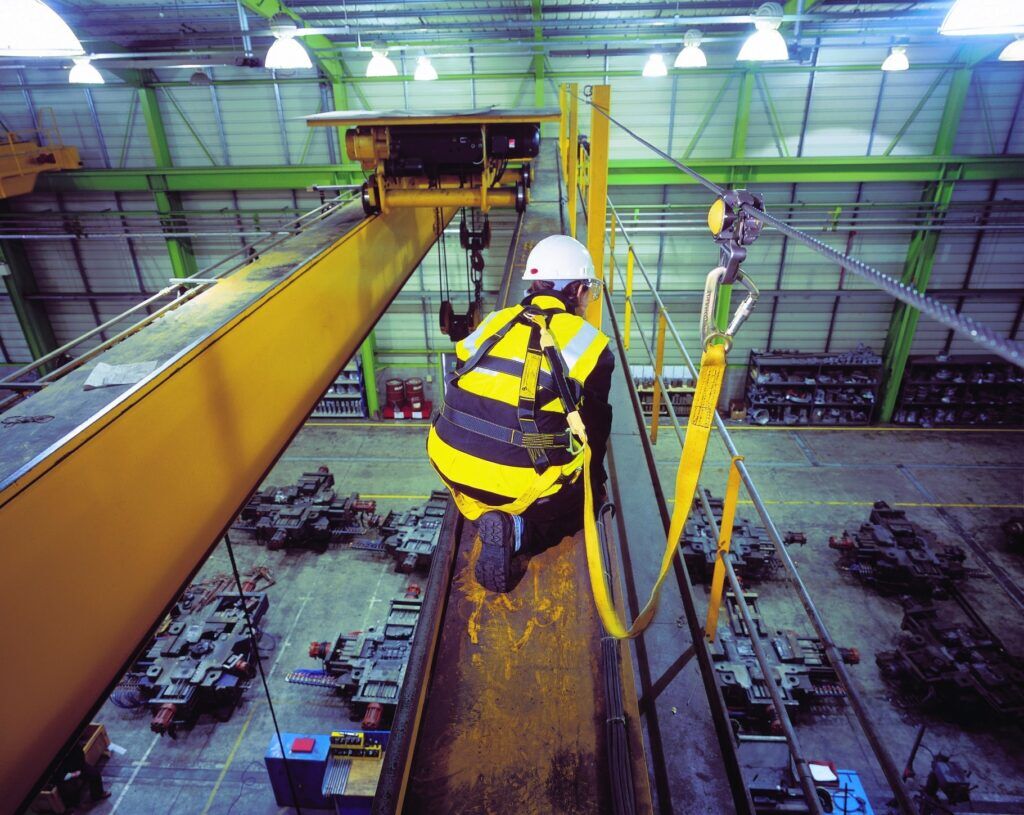 A worker in a safety harness is kneeling on a crane beam. They are showing fall protection measures in an industrial area. Machinery can be seen below them.